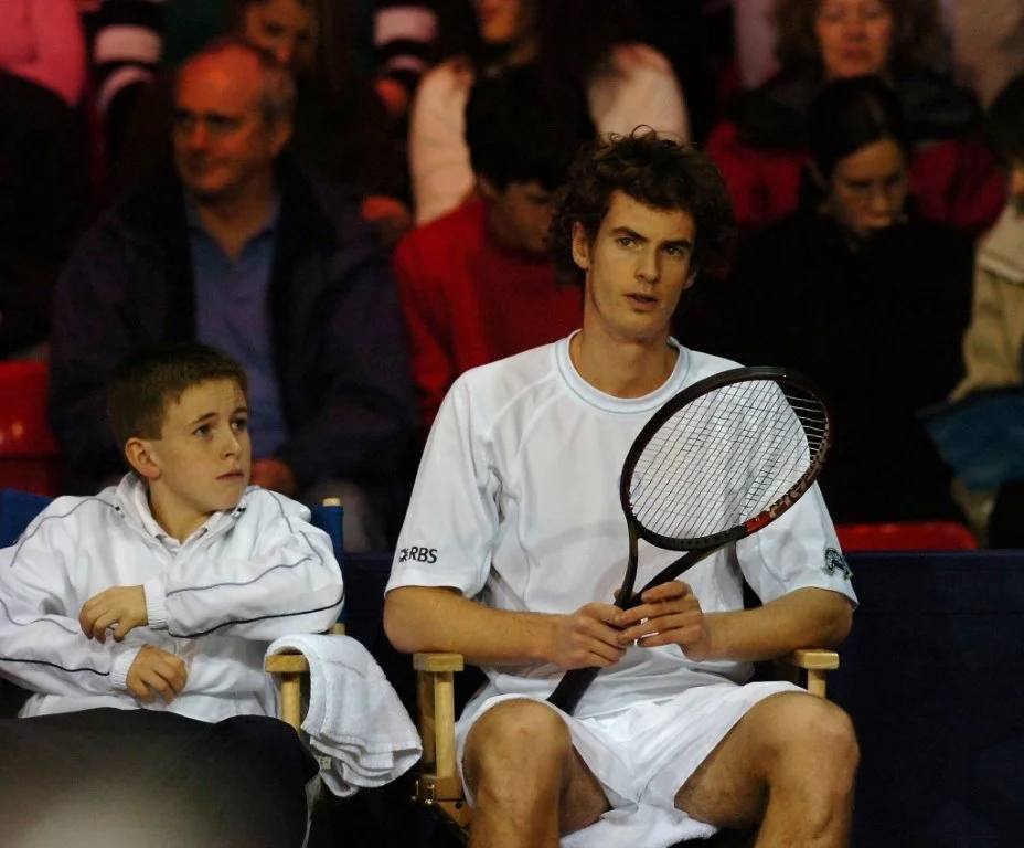 A professional tennis player seated courtside holding a racket, with a young player beside him